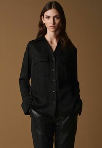 Sort satin skjorte med knapper og krave, to brystlommer og lange ærmer. Glat tekstur med en klassisk pasform. Parret med sorte læderbukser.