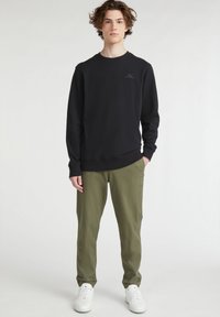 Sudadera negra con cuello redondo y puños, combinada con pantalones verde oliva; el modelo lleva zapatillas blancas y está de pie frente a un fondo neutro.