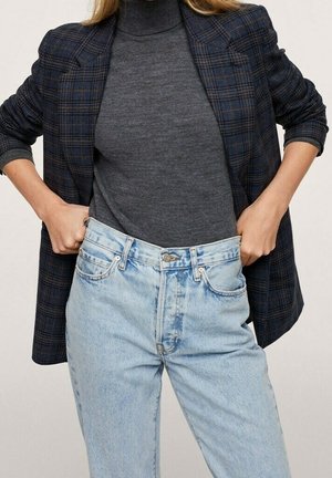 Femme portant un blazer à carreaux foncés sur un col roulé gris et un jean taille haute bleu clair, ajustant la taille avec les deux mains.