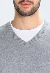 Pull en laine gris clair avec un col en V, présentant une texture lisse et des détails côtelés fins autour de l'encolure, porté sur une chemise blanche.