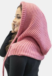 Sciarpa a maglia con cappuccio in un morbido rosa, con texture a coste, caratterizzata da una lunga coda e un design a strati. Indossata sopra una maglietta nera.