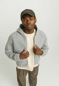 Grauer Fleece-Zipper-Hoodie mit Kapuze, über einem hellbeigen Pullover getragen. Das Model hat dunkelbraune Haut und trägt eine dunkle Mütze.