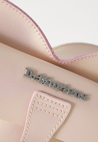Dr. Martens-veske i myk rosa syntetisk materiale, med sølvfargetlogo, kontrasterende rosa sømmer og en dekorativ brett øverst.