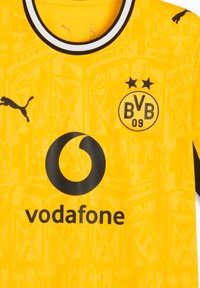Κίτρινη φανέλα ποδοσφαίρου της Borussia Dortmund με μαύρο λογότυπο Puma, χορηγό Vodafone, έμβλημα συλλόγου και διακριτικό γεωμετρικό σχέδιο φόντου.