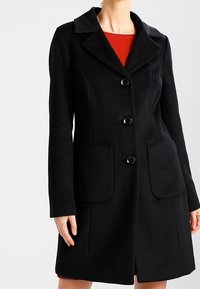 Zwarte wollen jas met een notch-kraag, vier knopen en twee voorkruiken. Licht getailleerde vorm, gladde textuur en minimalistisch design.