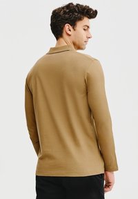 Brązowa, długos ławkowa koszulka polo o miękkiej fakturze, z kołnierzykiem i prostym krojem, noszona przez osobę odwróconą tyłem do aparatu.