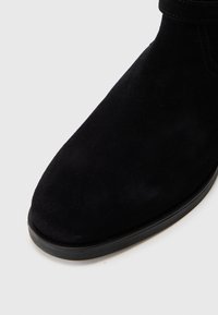 Czarne zamszowe botki za kostkę mają okrągły nosek, płaską gumową podeszwę oraz minimalne przeszycia, co nadaje im elegancki, czysty wygląd.