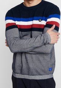 Marineblauer Rundhals-Pullover mit horizontalen roten, weißen und blauen Streifen. Gestrickte Textur, gerippte Bündchen und Saum, mit einem kleinen Logo-Patch.