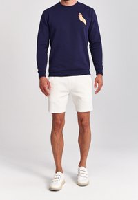 Felpa blu navy con maniche lunghe e scollo rotondo; presenta un design di un uccello leggermente colorato in rilievo sul petto, abbinata a pantaloni corti bianchi.