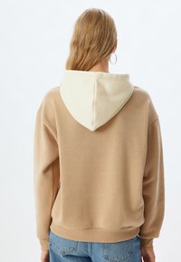 Sudadera beige con capucha crema, que presenta una textura suave, bolsillo canguro y puños acanalados. La parte trasera muestra un ajuste relajado y un dobladillo ligeramente caído.