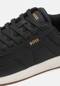 Mustade sneakers, mille tekstureeritud kangast ülemise osaga, millel on kuldne "BOSS" bränding, lame mustade paeltega ja valge kummist tallaga, millel on tekstureeritud muster.