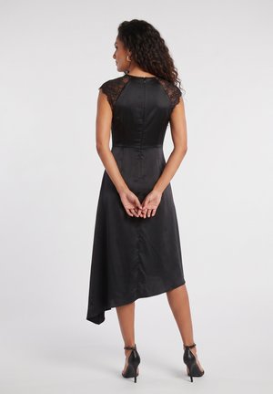 Femme aux cheveux bouclés de dos, portant une robe noire asymétrique avec des manches courtes en dentelle et des sandales noires à talons hauts.