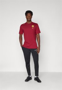 Punane lühikeste varrukatega T-särgi, millel on kollane logo, koos mustade kitsaste pükstega ja mustade spordijalatsitega, kannab seisev figuur.