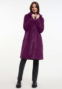 Abrigo de piel sintética morado con capucha, cierre de botones, dos bolsillos delanteros y corte relajado. Lleva puesto sobre pantalones oscuros y botas negras.