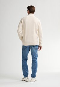 Maglione a maglia color crema con colletto alto e polsini a coste, abbinato a jeans in denim blu chiaro. Design semplice, materiale texturizzato.