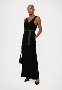 Mujer vestida con un maxi vestido de terciopelo negro sin mangas y con un lazo en la cintura, sosteniendo un bolso negro, de pie contra un fondo claro.