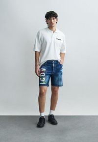 Blue denim shorts featuring colorful graphic detailing on the left leg, paired with a short-sleeve white polo shirt. Black sneakers.
