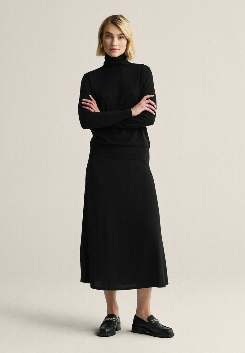 Nainen, jolla on lyhyet vaaleat hiukset, pukeutuneena mustaan pooloneuleeseen, mustaan midihameeseen ja mustiin loafer-kenkiin, joissa on kultainen ketjuyksityiskohta, seisoo käsivarret ristissä.