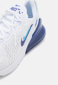 Vit atletisk sko med ovandel i mesh, blå detaljer och en synlig luftkuddsenhet i hälen. Har snören och perforerad design.