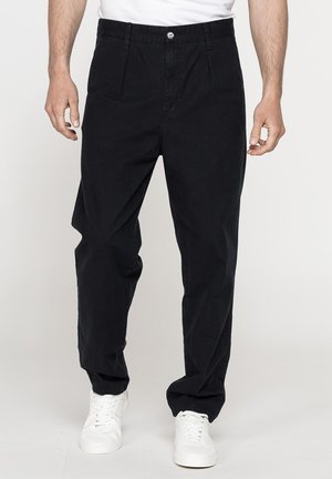 Uomo con pantaloni neri larghi e scarpe da ginnastica bianche in piedi davanti a uno sfondo neutro.