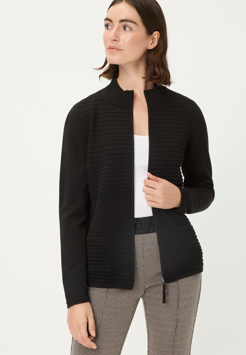 Schwarzer Cardigan mit strukturiertem Rippstrick, rundem Kragen und einem Reißverschluss vorne. Trägt man über einem weißen Oberteil und gemusterten Hosen.