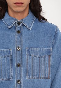 Denim shirt in light blue featuring a pointed collar, black buttons, two front pockets with vertical stitching, and a textured fabric.