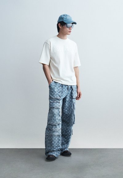 Jeune homme portant un t-shirt blanc avec un logo, un pantalon cargo bleu à motifs, une casquette de baseball bleue, des lunettes teintées et des chaussures noires, debout contre un mur uni.