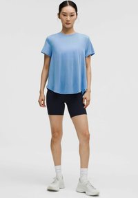Top athletic de culoare albastră cu mânecă scurtă și decolteu rotund, purtat cu pantaloni scurți negri cu talie înaltă. Modelul stă pe un fundal neutru.