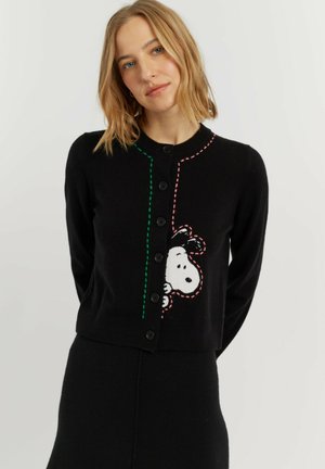Vrouw met blond haar die een zwarte gebuttonde cardigan draagt met een Snoopy cartoonfiguur die van opzij naar buiten gluurt, gecombineerd met een zwarte rok.