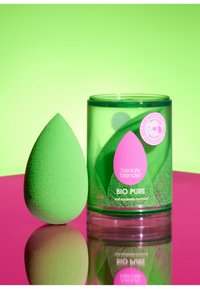 Spugna per il trucco verde con forma a goccia arrotondata, posizionata accanto a un contenitore cilindrico trasparente verde con dettagli e branding rosa.