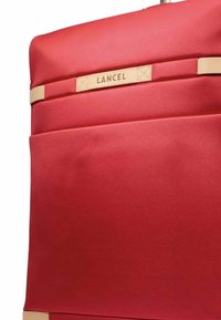 Coin de valise en tissu rouge avec des accents en cuir beige et une étiquette en cuir beige estampillée "LANCEL" sur fond blanc.