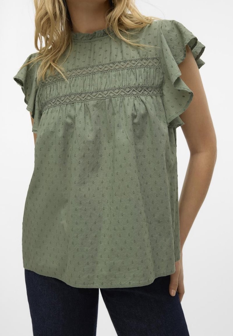 Blouse verte à manches courtes avec des pois texturés et des détails en dentelle, portée avec un jean bleu foncé sur une personne debout.