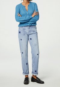 Jasnoniebieski kardigan z prążkowanej dzianiny z zapięciem na guziki, w połączeniu z jasnoniebieskimi dżinsami z haftem w niebieskie gwiazdki i podwiniętymi mankietami. Czarne loafersy z ćwiekami.