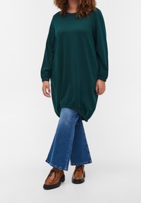 Leśnozielona, oversize'owa sukienka z długimi rękawami i zaokrąglonym hemem, zestawiona z niebieskimi dżinsami w stylu dzwonów i brązowymi butami na sznurówki.