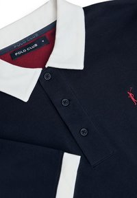 Marineblaues Poloshirt mit weißem Kragen, drei schwarzen Knöpfen, rotem Etikett innen und kleinem roten bestickten Logo auf der Brust. Strukturierter Stoff.
