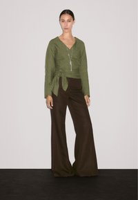 Un mannequin se tient devant un fond uni, portant un haut enveloppant vert et un pantalon large marron, avec un collier en pendentif argenté.