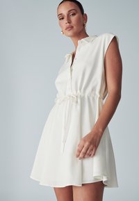 Vestido corto blanco con cuello tipo polo, cierre frontal de botones y cintura con lazo elástico. Presenta mangas cortas y una falda fluida.