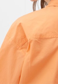 Orange skjorte med en afslappet pasform, letvægtigt stof og et simpelt design. Bagsiden præsenterer en diskret krave og syningsdetaljer.