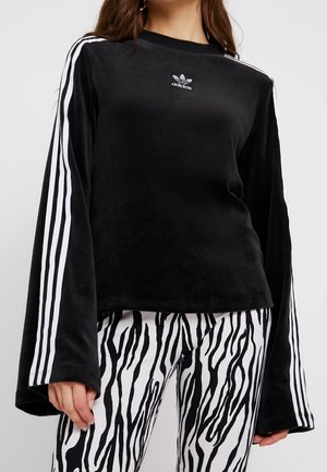Kobieta ubrana w czarny długi rękaw Adidas z białymi paskami na rękawach i czarno-białe spodnie z nadrukiem zebry, stojąca na jednolitym tle.