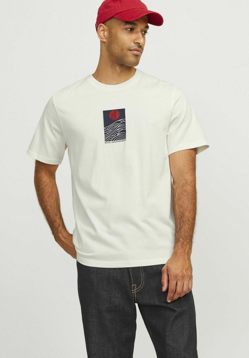 White cotton t-shirt featuring a graphic print of waves and a red sun. Paired with dark denim jeans. Simple round neckline and short sleeves.
