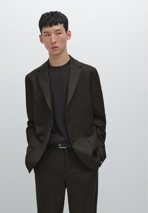 Dark brown tailored suit jacket with notched lapels, worn over a black shirt. Pants match the jacket, with a slim black belt featuring a silver buckle.