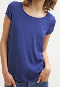 Top blu a maniche corte in tessuto liscio, caratterizzato da una scollatura rotonda e una piccola tasca sul petto. Design semplice, vestibilità morbida e senza decorazioni.