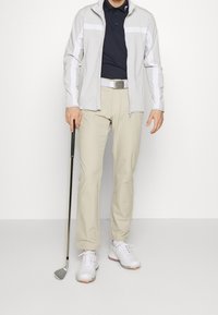 Vaaleanharmaa vetoketjullinen takki, jossa valkoiset raidat, tummansininen poolopaita, beiget housut ja valkoiset golfkengät. Pitelee golfmailaa.