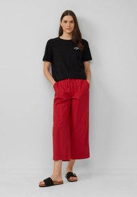 Zwarte katoenen t-shirt met korte mouwen en wit logo, gecombineerd met losvallende rode cropped broeken en zwarte slide sandalen.