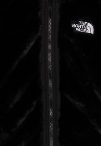 Chaqueta negra y esponjosa hecha de suave piel sintética, con cremallera frontal y logotipo bordado en el pecho izquierdo. Textura suave.