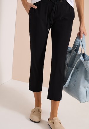 Persona con pantalones cortos negros y zapatos beige sin cordones, sosteniendo un bolso tote de mezclilla azul claro en una mano.