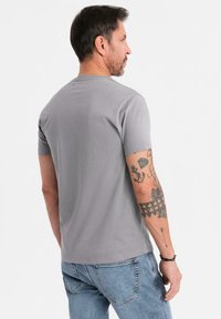 Szary T-shirt z krótkim rękawem i okrągłym dekoltem, wyeksponowany z tyłu, zestawiony z jasnoniebieskimi dżinsami, z widocznymi tatuażami na ramieniu.