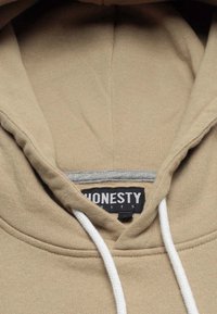 Beige Kapuzenpullover mit grauem Innenfutter, weißen Kordeln und einem schwarzen Etikett mit der Aufschrift "HONESTY", das in Weiß gestickt ist.