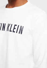 Osoba v bílém svetru s velkým černým nápisem "CALVIN KLEIN" na hrudi.