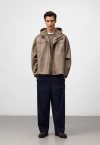 Brązowa skórzana kurtka z kapturem i polarowym podszyciem, noszona na szarym swetrze i granatowych szerokich spodniach. Gładka tekstura, luźny fason.